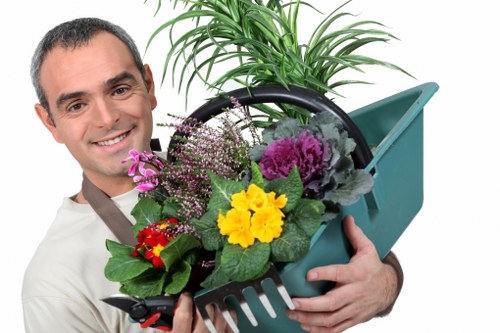The image depicts a smiling man holding a teal plastic garden tool carrier filled with a variety of potted plants, including flowering and foliage plants with green leaves and vibrant blooms. Behind him, there is a garden scene with neatly maintained lawn grass, showing dense, healthy green turf typical of suburban gardens in Upminster. The background features a large shrub or small tree, along with a paved or patio area that appears well-kept and suitable for outdoor activities. The overall scene suggests regular gardening and lawn care, consistent with professional services offered by Lawn Mowing Upminster, with the environment illuminated by natural daylight under overcast weather conditions, highlighting the lush greenery and vibrant plant colours in a tidy outdoor space.